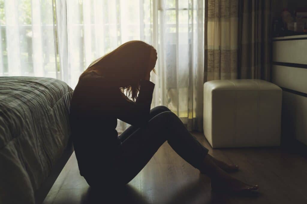 Woman sitting on the floor next to a bed with her head in her hands